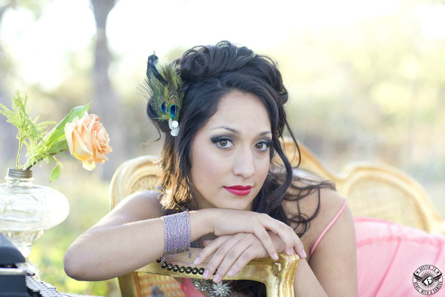 brunette latino fashion model with curly hair and peacock feather headpiece with brown eyes and red lips in a pick dress and chain wristlet sits in a vintage gold sofa in the fall outdoors with a rose in antique lamp holder    